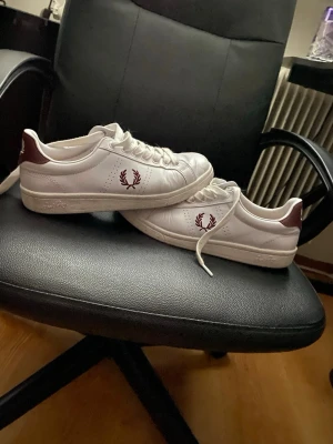 Vita Fred Perry sneakers med vinröda detaljer - Snygga vita sneakers från Fred Perry med vinröda detaljer och logga på sidan. Skorna har klassisk låg profil, vita snören och är gjorda i läder. Perfekta för en clean och stilren look.