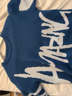 Blå stickad tröja med vit text - Cool blå stickad tröja med vit grafisk text över framsidan. Tröjan har rund halsringning och långa ärmar. Perfekt för dig som gillar streetwear och vill sticka ut med en unik design.