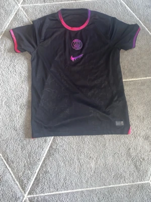 Svart PSG Mbappé tröja Nike - Snygg svart PSG matchtröja från Nike med rosa och lila detaljer. Framsidan har PSG-logga och Nike-logga i rosa, baksidan har Mbappé och nummer 7 tryckt i vitt och rosa. Korta ärmar och rund hals. Perfekt för fotbollsfans!
