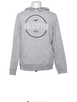 Grå hoodie från Hollister - Snygg trendig grå hoodie från Hollister med tryck framtill och texten 'Hollister 1922'. Klassisk känguruficka, dragsko i huvan och ribbade muddar. Perfekt för en avslappnad och trendig stil. Mjuk bomullsblandning för extra komfort.