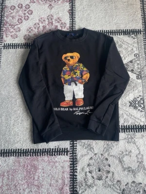 Svart Polo Bear sweatshirt Ralph Lauren - Svart sweatshirt från Ralph Lauren med ikoniskt Polo Bear-tryck framtill. Björnen har på sig en färgglad hawaiiskjorta, vita byxor och solglasögon. Tröjan har rund hals och ribbade muddar. Perfekt för en avslappnad och cool stil. Påse ingår och pris kan diskuteras vid snabb affär.