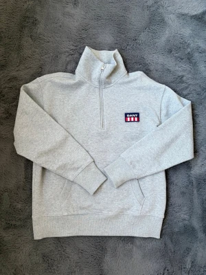 Grå halvzip från GANT - Snygg grå halvzip hoodie från GANT med broderad logga på bröstet i blått, vitt och rött. Tröjan har hög krage, känguruficka och ribbade muddar. Perfekt för en chill och stilren look. Mjuk bomullsblandning som känns skön mot huden. Otroligt fin och använd endast EN gång. Skriv vid frågor eller funderingar. 
