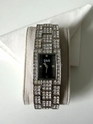 D&G silverfärgad klocka med stenar - Snygg klocka från D&G i silverfärg med rektangulär svart urtavla och armband täckt av glittrande stenar. Osäker på om den är äkta därav pris. Batteri behöver bytas 🤍 extra bitar medföljer 