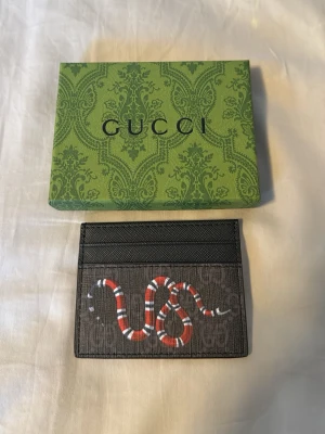 Gucci korthållare - Snygg korthållare från Gucci i svart canvas med klassiskt GG-mönster och en rödvit orm på framsidan. Plånboken har flera kortfack och är perfekt för dig som vill ha något stilrent men ändå sticker ut. Kommer med originalask.