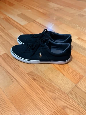 Svarta Polo Ralph Lauren sneakers - Svarta sneakers från Polo Ralph Lauren med klassisk låg profil och vit sula. Skorna har snörning och den ikoniska guldiga Polo-loggan broderad på sidan. Tillverkade i textilmaterial med bekväm innersula. Perfekta för en stilren och avslappnad look.