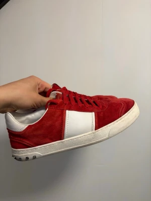 Valentino flycrews röda sneakers  - Riktigt snygga röda sneakers från Valentino Garavani i mocka med vita detaljer och platt sula. Skorna har en bred vit panel på sidan, vita partier vid häl och plös samt röda skosnören. Ikoniska nitar baktill på sulan ger extra edge.