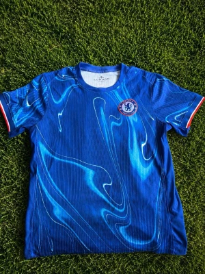 Chelsea FC blå matchtröja - Snygg blå Chelsea FC matchtröja med unikt mönster i olika blå nyanser och klubbens emblem på bröstet. Tröjan har korta ärmar med röda och vita detaljer vid ärmsluten. Perfekt för fotbollsträning eller att bära på läktaren.