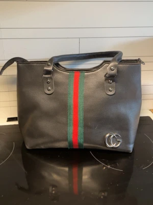 Svart handväska med röd och grön rand - Snygg svart handväska i läder med klassiska röda och gröna ränder framtill. Väskan har dubbla handtag och silverfärgad GG-logga i metall på framsidan. Perfekt för dig som vill ha en stilren och trendig accessoar.