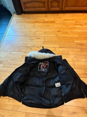 Moose knuckles jacka - Snygg svart dunjacka från Moose Knuckles med pälskantad huva och quiltad design. Jackan har dragkedjor vid ärmslut och en stor logga på insidan. Perfekt för kalla vinterdagar och riktigt varm tack vare dunfyllningen.