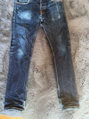 Mörkblå slim fit jeans från Nudie - Snygga mörkblå slim fit jeans från Nudie med klassiska fem fickor och slitna detaljer på knän och lår. Jeansen har kontrastsömmar och en något tvättad look. Perfekta för en avslappnad och trendig stil. Storlek 30 34