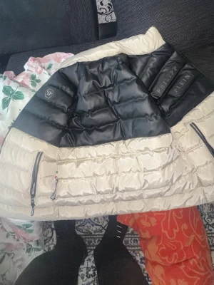 Philipp Plein svart och beige pufferjacka - Snygg pufferjacka från Philipp Plein i svart och beige med märkets logga på bröstet och detaljerade dragkedjor. Jackan har en modern, boxig passform och är perfekt för kalla vinterdagar. Materialet är glansigt och ger en exklusiv känsla.