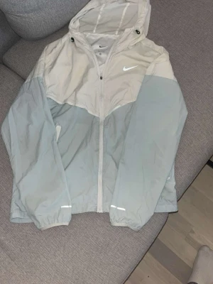 Nike vindjacka i vitt och ljusblått - Snygg vindjacka från Nike i vitt och ljusblått med dragkedja framtill och huva. Jackan har elastiska muddar vid ärmsluten och reflekterande detaljer på ärmarna. Perfekt för blåsiga dagar och sportiga outfits. Bra till sommaren. 