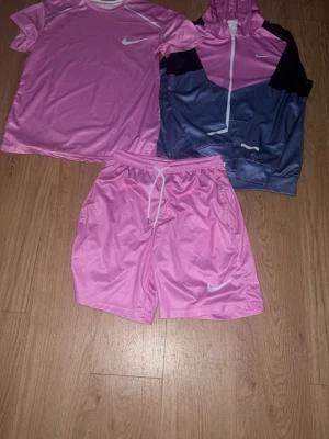Nike miller Rosa - Säljer ett par snygga rosa sportshorts samt t-shirt och kofta från Nike med vit snörning i midjan och klassisk logga på benet. Shortsen är tillverkade i ett glansigt syntetmaterial och har en avslappnad passform, perfekta för träning eller chill.
