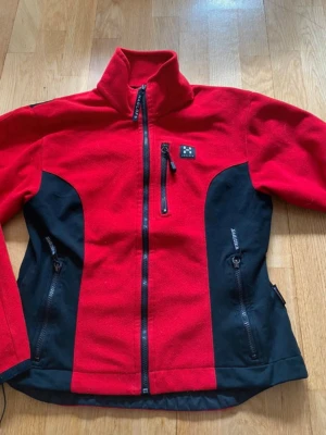 Röd och svart fleecejacka Haglöfs - Snygg röd och svart fleecejacka från Haglöfs med hög krage och tre dragkedjeförsedda fickor. Perfekt för kyliga dagar och outdoor-aktiviteter. Jackan har en sportig look och är tillverkad i mjuk fleece med svarta detaljer på sidorna. Strl L men mer S/m