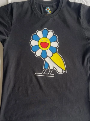 Svart t-shirt med färgglad blomfågel - Cool svart t-shirt från OVO med en stor, färgglad blomfågel på bröstet i blått, vitt, gult och rött. T-shirten har rund hals och korta ärmar. Perfekt för dig som gillar unika och lekfulla prints.