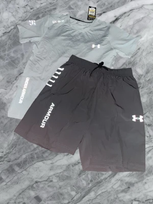 Under Armour träningsset grå/mörkgrå - Snyggt träningsset från Under Armour med en ljusgrå t-shirt och svarta shorts. T-shirten har tryckt logga och text, medan shortsen har dragsko i midjan och reflektiva detaljer. Perfekt för gymmet eller löprundan. OBS!! Köp inom 48 timmar få 149kr rabatt!!