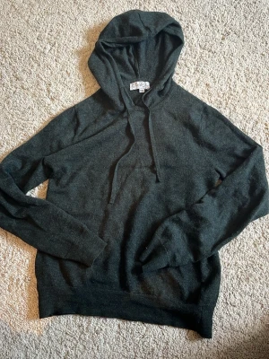 Mörkgrå hoodie i mjukt material - En stilren mörkgrön hoodie från Ahlquist Clothing, ett mjukt och skönt material. Klassisk modell med huva och dragsnören, långa ärmar och ribbade muddar vid ärmslut och nederkant. Perfekt för en avslappnad och trendig look.