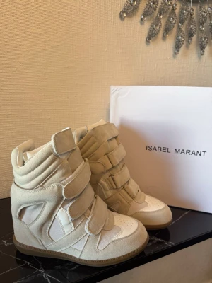 Isabel Marant höga  - Snygga höga sneakers från Isabel Marant i beige mocka och vitt läder. Skorna har flera kardborreband, vadderad plös och häl samt en gummisula med mönster. Perfekta för dig som gillar streetstyle och vill ha något unikt till din outfit.