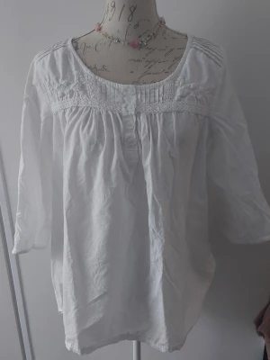 Vit blus med spets och broderi - Söt vit blus med trekvartsärm, dekorativa knappar framtill och fina spets- och broderidetaljer upptill. Blusen har rund halsringning och lätt rynkade partier för en avslappnad look. 