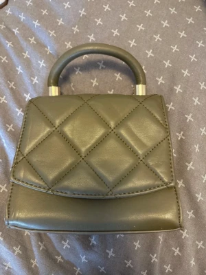 Olivgrön quiltad handväska - Snygg olivgrön handväska i quiltat mönster med stilrent handtag och silvriga detaljer. Väskan har en boxig form och är tillverkad i konstläder. Perfekt accessoar för att lyfta din outfit.