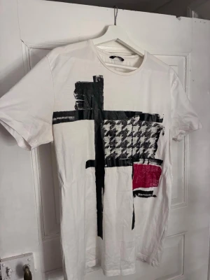 Vit t-shirt från Antony Morato - Vit t-shirt från Antony Morato med coolt grafiskt tryck i svart, vitt och rött framtill. T-shirten har rund halsringning och korta ärmar. Materialet ser ut att vara mjuk bomull, perfekt för en trendig och avslappnad look.