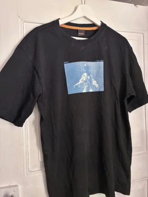 Svart BOSS t-shirt med astronauttryck - Cool svart t-shirt från BOSS med ett blått tryck av en astronaut på bröstet och texten 'TEST 001'. T-shirten har rund hals och korta ärmar. Perfekt för dig som gillar streetwear och vill sticka ut med en unik print.