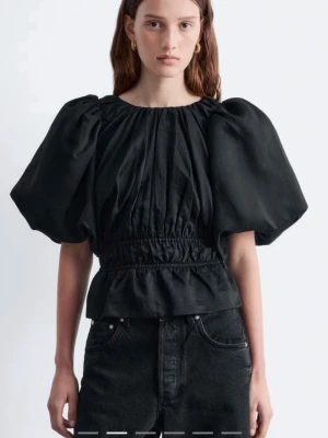 Svart blus med puffärmar - Trendig svart blus med stora puffärmar och markerad midja. Blusen har rund halsringning och volang nedtill, vilket ger en snygg siluett. Perfekt att styla med jeans eller kjol för en cool look.