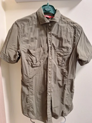 Fjällräven khaki, kortärmad skjorta - Snygg beige skjorta med korta ärmar och två bröstfickor. Skjortan har klassisk krage och knäppning framtill. Ultralätt, snabbtorkande. Perfekt på resan för en avslappnad stil och enkel att matcha med jeans eller shorts.