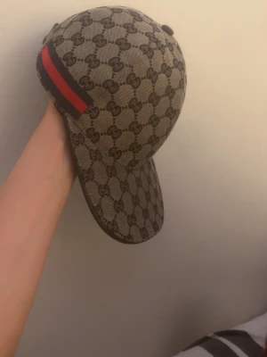 Gucci keps med GG-mönster - Snygg keps från Gucci i beige med klassiskt GG-monogram över hela kepsen. Baktill finns en ikonisk röd och marinblå rand. Kepsen har böjd skärm och är tillverkad i canvasmaterial. Perfekt för dig som vill sticka ut med en lyxig accessoar.