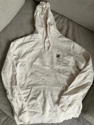 Vit hoodie från Lyle & Scott - Snygg vit hoodie från Lyle & Scott med klassisk gul logga på bröstet. Hoodien har huva med dragsko, magficka och ribbade muddar vid ärmslut och nederkant. Perfekt för en chill och stilren look.