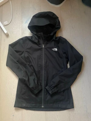 Svart regnjacka The North Face - Svart regnjacka från The North Face med huva och hel dragkedja framtill. Jackan är tillverkad i vattentätt DryVent-material och har elastiska muddar vid ärmsluten. Logga på bröstet och DryVent-tryck på ärmen. Perfekt för regniga dagar.