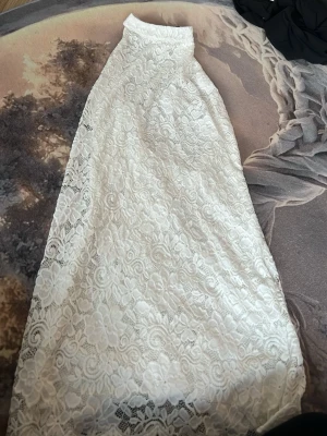 Vit lång spetsklänning - Elegant vit långklänning med vacker spets och blommigt mönster. Klänningen har hög hals och är ärmlös, vilket ger en stilren och romantisk look. Perfekt för sommarens speciella tillfällen eller studentfirande.