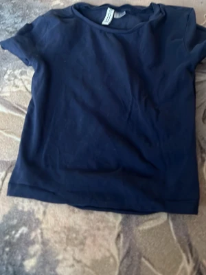 Mörkblå croppad basic t-shirt från H&M Divided - En enkel och stilren mörkblå t-shirt från H&M Divided Basic. T-shirten har korta ärmar och rund halsringning. Perfekt till jeans eller shorts för en avslappnad look. Tillverkad i mjuk bomull för skön komfort.