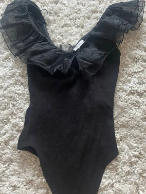Svart body med volang från AMISU - Snygg svart body från MISU med stora volanger i mesh runt urringningen. Kroppsnära passform och djup v-ringning. Perfekt för dig som vill sticka ut med en unik detalj. Materialet är stretchigt och bekvämt.