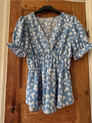 Blommig blå blus med puffärm - Ljusblå blus med vita blommor, v-ringning och puffiga kortärmar med volang och knyt. Smockad midja ger en fin siluett och blusen är gjord i ett lätt och luftigt material, perfekt för sommar och vår.