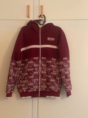 Vinröd hoodie från BOSS med tryck - Snygg vinröd hoodie från BOSS med vit dragkedja och huva. Framsidan har en vit rand över bröstet och nedre delen är täckt av vitt BOSS-tryck. Muddar vid ärmslut och nederkant. Perfekt för dig som vill sticka ut med en streetstyle-look.