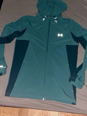 Grön vindjacka från Under Armour - Snygg grön vindjacka från Under Armour med huva och hel dragkedja. Jackan har reflekterande detaljer, elastiska muddar och två sidofickor. Perfekt för blåsiga dagar och sportiga aktiviteter.