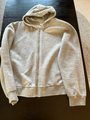 Gina Tricot - Grå zip hoodie med huva - En enkel och stilren grå hoodie med dragkedja framtill och huva med dragsko. Tillverkad i mjukt sweatshirtmaterial och har ribbade muddar vid ärmar och nederkant. Perfekt för en avslappnad och bekväm look.