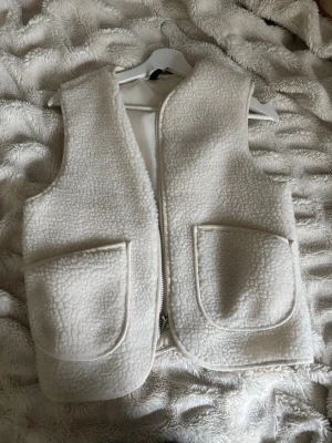 Vit teddyväst med dragkedja - Mysig vit teddyväst med två stora fickor framtill och dragkedja. Västen har en fluffig och mjuk yta i fuskpälsliknande material som ger en trendig look. Perfekt att slänga över en hoodie eller tröja för extra värme.