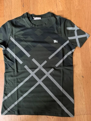 Grön Burberry t-shirt med mönster - Snygg grön t-shirt från Burberry med klassiskt rutmönster i svart och vitt. T-shirten har rund hals, korta ärmar och en liten broderad Burberry-logga på bröstet. Perfekt för dig som gillar stilrena och trendiga plagg.