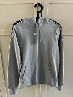 Grå Nike hoodie med svarta detaljer - Snygg grå hoodie från Nike med huva och dragsko. Har svarta paneler med vita Nike-loggor längs ärmarna och en klassisk känguruficka framtill. Perfekt för en sportig och avslappnad stil. Materialet är mjukt och bekvämt bomullsblandat tyg.