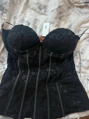 Svart spetskorsett från Zara - Snygg svart korsett från Zara med tunna axelband och vadderade kupor. Korsetten är täckt av blommig spets och har markerade sömmar framtill för en klassisk siluett. Insidan är beige för en snygg kontrast. Perfekt för att lyfta din outfit.