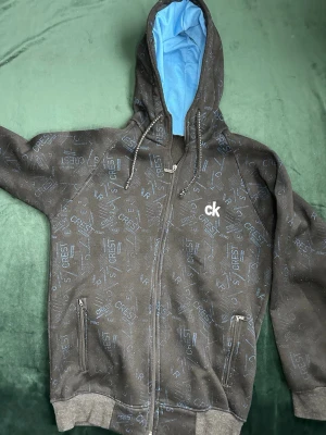 Svart CREST hoodie med dragkedja - Snygg svart hoodie från CREST med dragkedja framtill och två fickor med dragkedja. Hoodien har ett diskret mönster med CREST-loggor i blått och justerbar huva med snören. Insidan av huvan är ljusblå för en cool kontrast. Perfekt för en avslappnad streetwear-look. Pris kan diskuteras. MVH stentorparchive⚓️