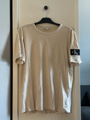 Beige t-shirt från Calvin Klein - Snygg beige t-shirt från Calvin Klein med klassisk passform och korta ärmar. T-shirten har en svart patch med CK Jeans-logga på vänster ärm och är gjord i mjuk bomull. Perfekt till jeans eller shorts för en clean och enkel look.