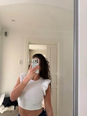 Vit croppad topp med volangärmar - Säljer en vit croppad topp med volangdetaljer vid axlarna. Toppen är ärmlös och har en rund halsringning. Perfekt för sommarens alla tillfällen och enkel att styla med jeans eller kjol.