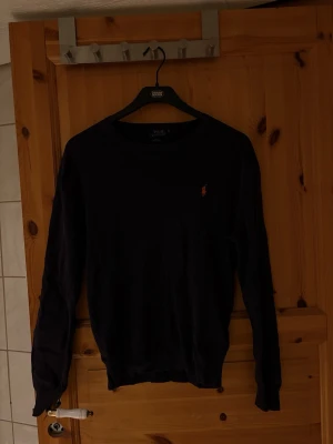 Svart tröja från Polo Ralph Lauren - Klassisk svart tröja från Polo Ralph Lauren med rund halsringning och den ikoniska broderade loggan i orange på bröstet. Långärmad modell i mjukt material, perfekt för en stilren look.