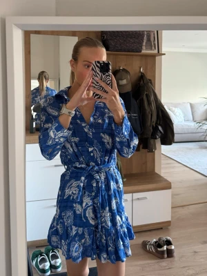 Blå kortklänning med blommigt mönster från Zara - Snygg blå kortklänning med vitt blommigt mönster och volangkant. Klänningen har lång ärm, knappar framtill och ett matchande knytbälte i midjan. Perfekt för dig som gillar en feminin och fräsch stil. Från Zara i storlek XS