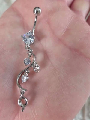 Silvrigt navelpiercingsmycke med stenar - Snyggt navelpiercingsmycke i silverfärgad metall med flera glittrande, klara stenar. Designen är böjd med hängande stenar i olika storlekar som ger ett elegant och trendigt intryck. Perfekt accessoar för att lyfta din stil.