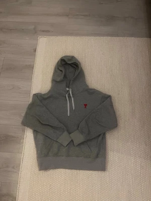 Grå hoodie från Ami Paris - Säljer en grå hoodie från Ami Paris med huva och dragsko. Klassisk passform med ribbade muddar och en liten röd Ami-logo broderad på bröstet. Tillverkad i mjuk bomullsblandning som är perfekt för chill dagar. Fråga om du undrar något!