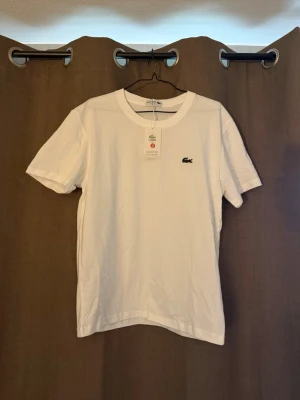 Vit t-shirt från Lacoste - Klassisk vit t-shirt från Lacoste med korta ärmar och rund halsringning. T-shirten har den ikoniska krokodilloggan broderad på bröstet och är tillverkad i mjuk bomull. Perfekt för en stilren och enkel look.